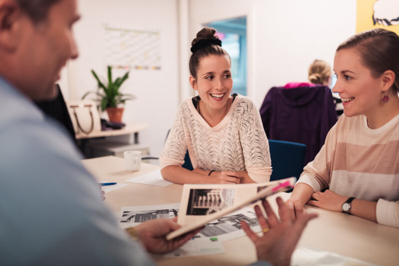 Zwei junge Frauen lächeln bei einem Gespräch am Tisch mit Informationsmaterialien in der Hand