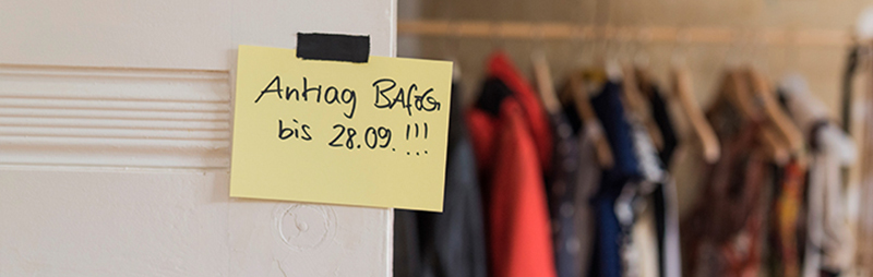 An einer weißen Tür klebt ein gelber Klebezettel mit der Aufschrift „Antrag BAföG bis 28.09.!!!“ Im Hintergrund hängen Kleidungsstücke leicht unscharf an einer Kleiderstange.
