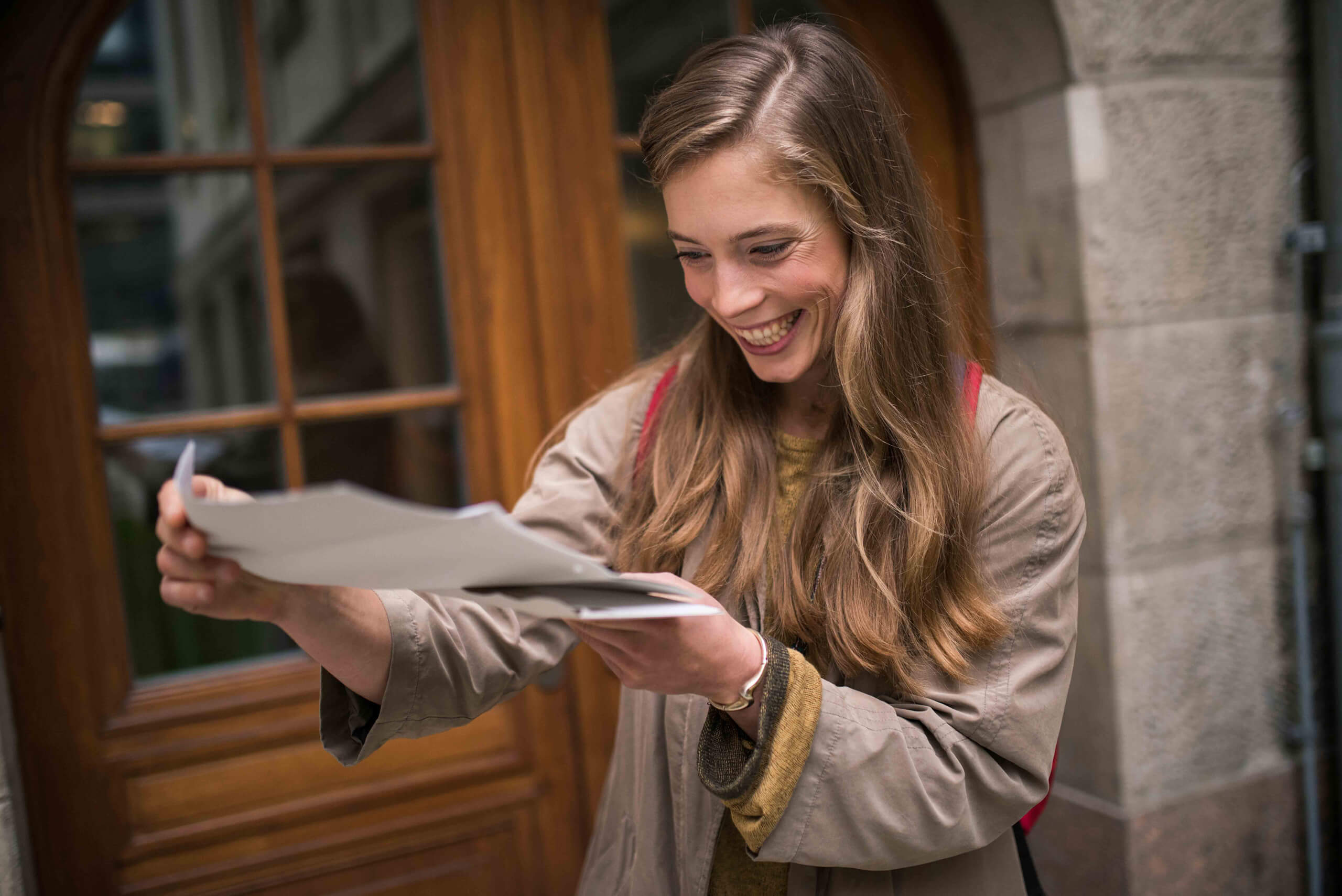 Junge Frau mit langen Haaren freut sich sichtlich über offizielle Dokumente, die sie draußen vor einem Holztor durchliest
