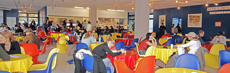 Buntes Café mit roten, gelben und blauen Stühlen, in dem Studierende an runden Tischen sitzen und essen oder sich unterhalten.
