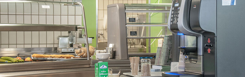 Detailansicht der Essensausgabe eines Cafés mit modernen Geräten und bereitgestelltem Kaffeeautomaten im Vordergrund.