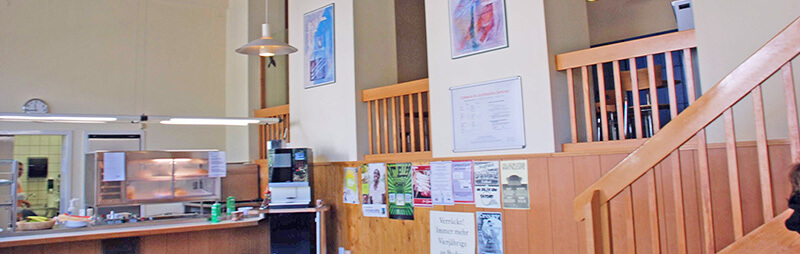 Innenansicht eines Cafés mit Holzverkleidung und Galerie, an der Theke steht eine Person neben einem Kaffeeautomaten.