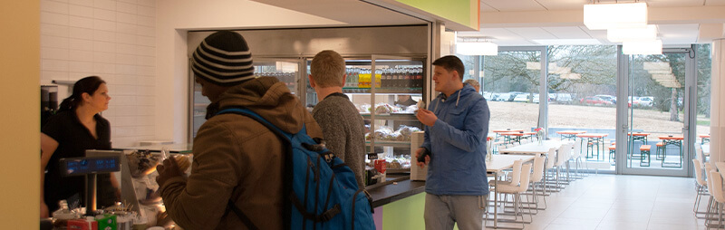 Mehrere Studierende stehen an der Essensausgabe eines modernen Campus-Cafés, während eine Mitarbeiterin bedient.