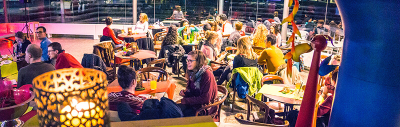 Menschen sitzen in einem lebhaften, bunt beleuchteten Café oder Restaurant mit lockerer Atmosphäre und moderner Einrichtung.