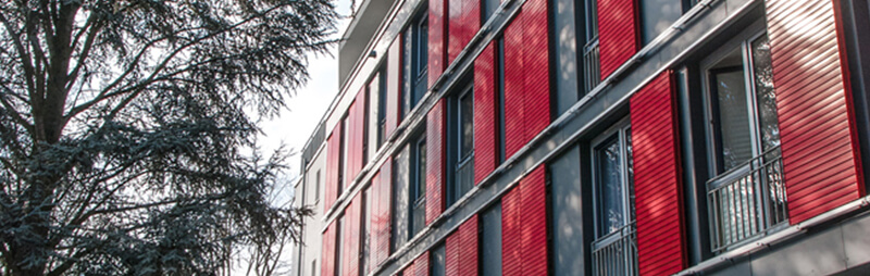 Modernes Wohngebäude mit roten Fassadenelementen, bodentiefen Fenstern und Schattenwurf durch umliegende Bäume