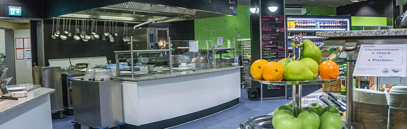 Moderne Cafeteria mit Edelstahl-Theke, offenem Küchenbereich, frischem Obst im Vordergrund und Getränkekühlschränken im Hintergrund.