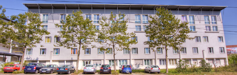 Großes Wohnheim mit flacher Dachkonstruktion, breiter Fensterfront und vorgelagerter Baumreihe sowie parkenden Autos davor