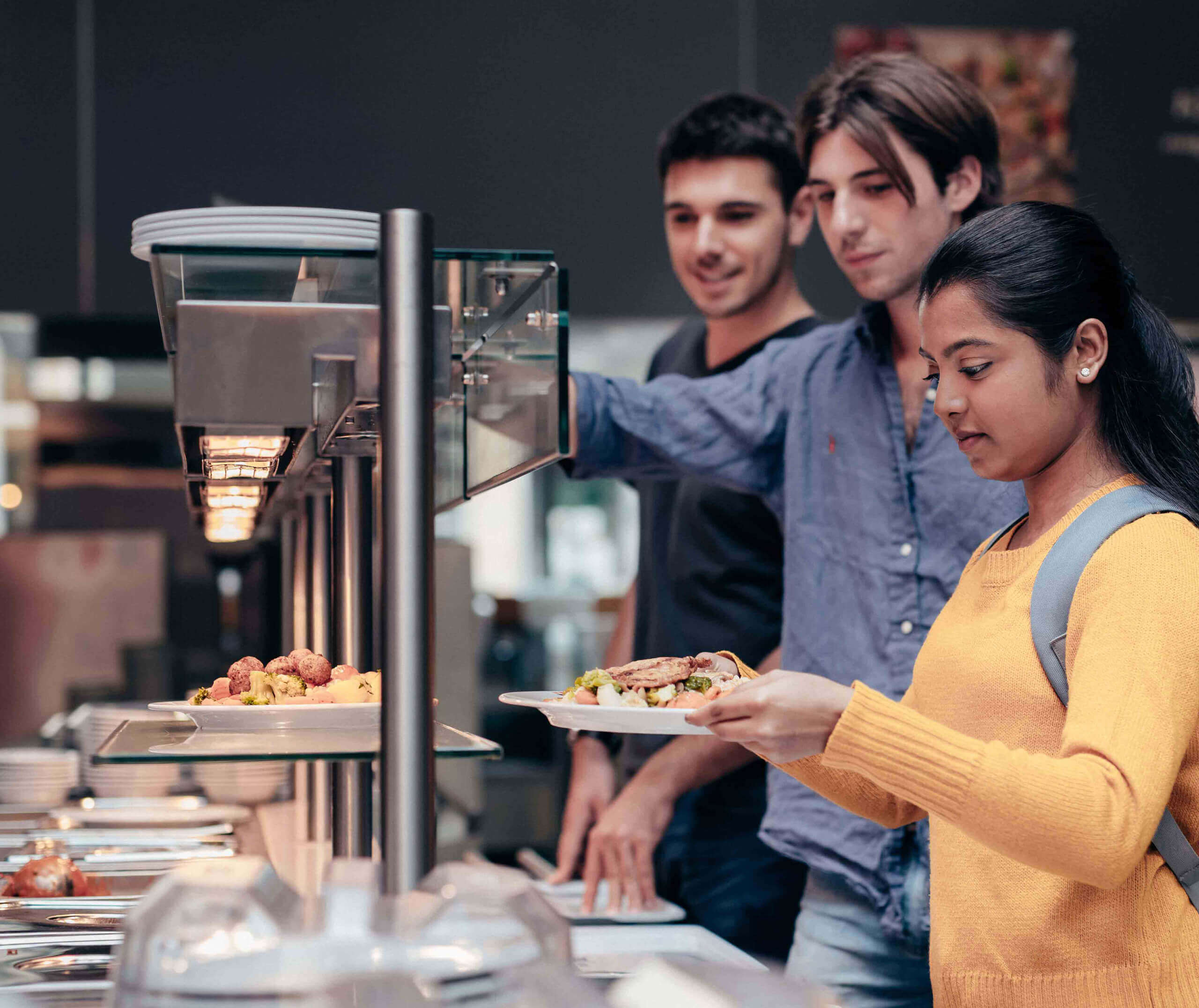 Drei junge Erwachsene stehen in einer Cafeteria Schlange und bedienen sich am Buffet. Die Frau vor ihnen, in einem gelben Pullover, hält einen Teller in der Hand und schaut sich das Angebot an.