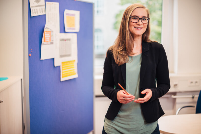Lächelnde Frau im Blazer steht vor einer Pinnwand mit Plänen und Informationen im Büro