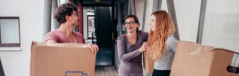 Drei Studierende tragen Umzugskartons auf einem offenen Laubengang eines Wohnheims und unterhalten sich lachend