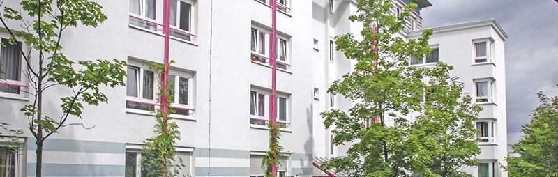 Modernisiertes Wohnheim mit heller Putzfassade, pinken Fassadenelementen und Bäumen vor dem Gebäude