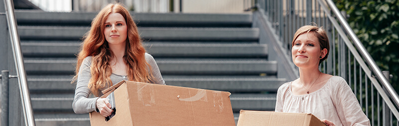 Zwei junge Frauen tragen Pappkartons eine Außentreppe hinauf, im Hintergrund sind Metallgeländer und Grünpflanzen zu sehen.