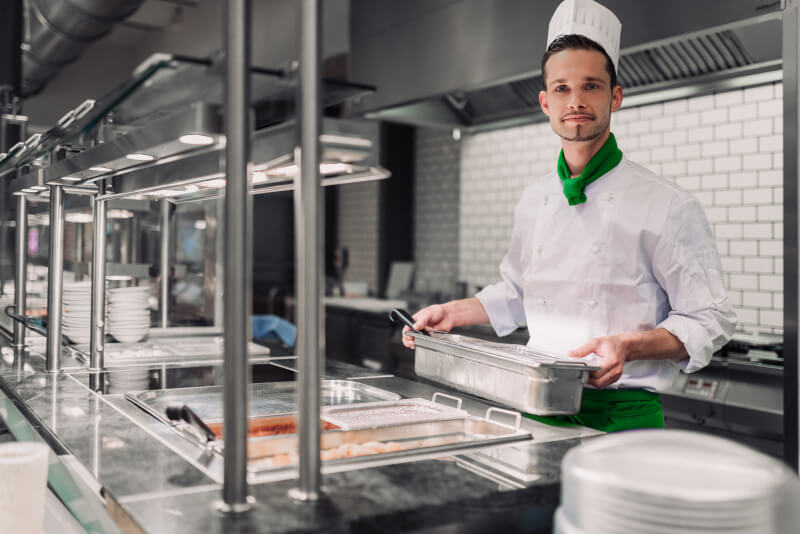 Koch in weißer Uniform mit grünem Halstuch an der Essensausgabe einer modernen Mensa