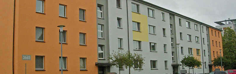 Mehrteiliger Wohnheimkomplex mit farblich abgesetzten Fassaden in Orange, Grau und Gelb, symmetrisch angeordneten Fenstern und kleinen Bäumen entlang der Straße.