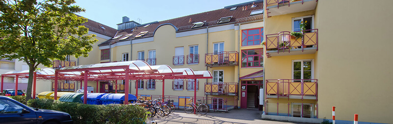 Gelb gestrichenes Wohnheim mit roten Balkonen, überdachten Fahrradstellplätzen und Recycling-Containern im Vordergrund