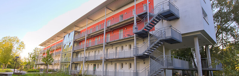 Vierstöckiges Wohnheim mit außenliegenden Fluren und Treppen vor roter Fassade im Grünen