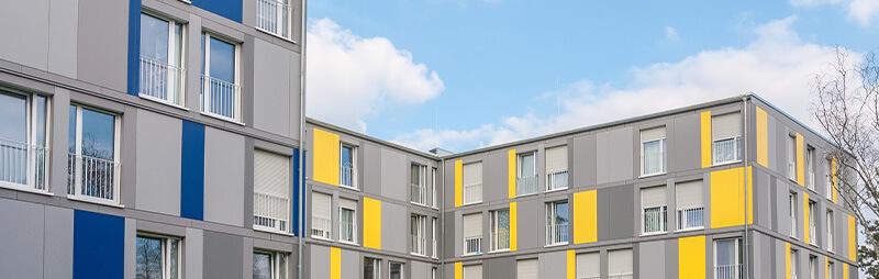 Wohnheim mit moderner grauer Fassade und senkrechten Farbstreifen in Gelb und Blau bei bewölktem Himmel