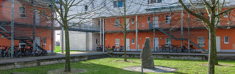 Innenhof eines Wohnheims mit roter Fassade, Außenfluren und abgestellten Fahrrädern neben einem Baum mit Skulptur