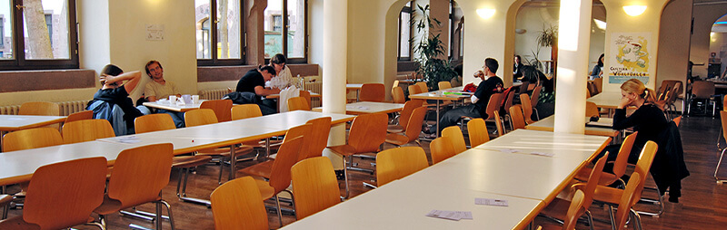 Großer heller Speisesaal mit langen weißen Tischen und vielen Holzstühlen, vereinzelt besetzt von Personen beim Essen oder Unterhalten.