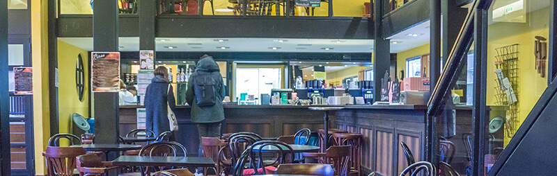 Gemütliches Café mit dunklen Holzmöbeln auf zwei Ebenen, zwei Personen stehen an der Theke, während andere sitzen oder sich unterhalten.