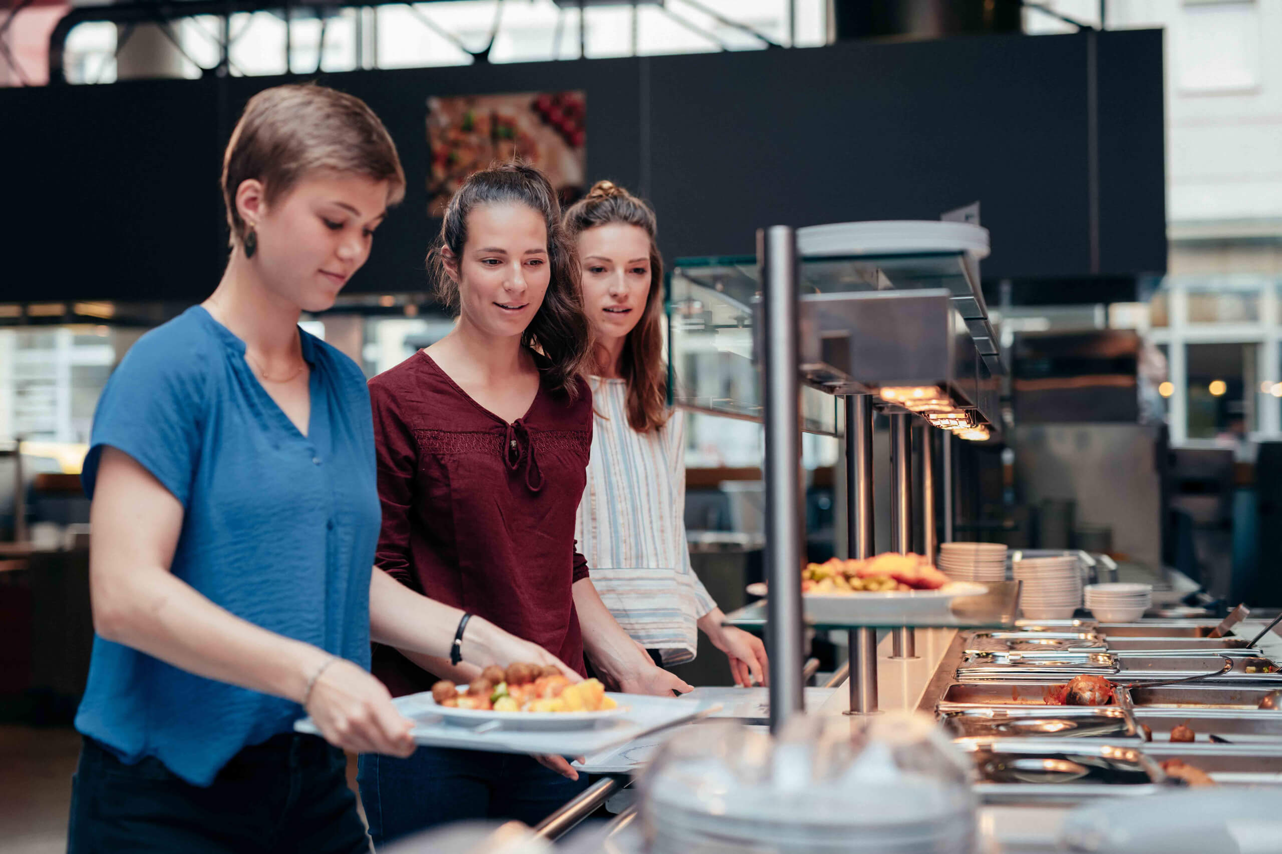 Drei junge Frauen stehen an einer Essenstheke im Cafeteria-Stil Schlange und bedienen sich unter warmem Licht mit warmen Speisen auf Tellern. In den Regalen finden sich verschiedene Gerichte, und das Ambiente wirkt modern und leger.
