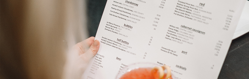 Eine Person betrachtet eine umfangreiche Getränkekarte, im Vordergrund ist ein Glas mit Cocktail sichtbar.