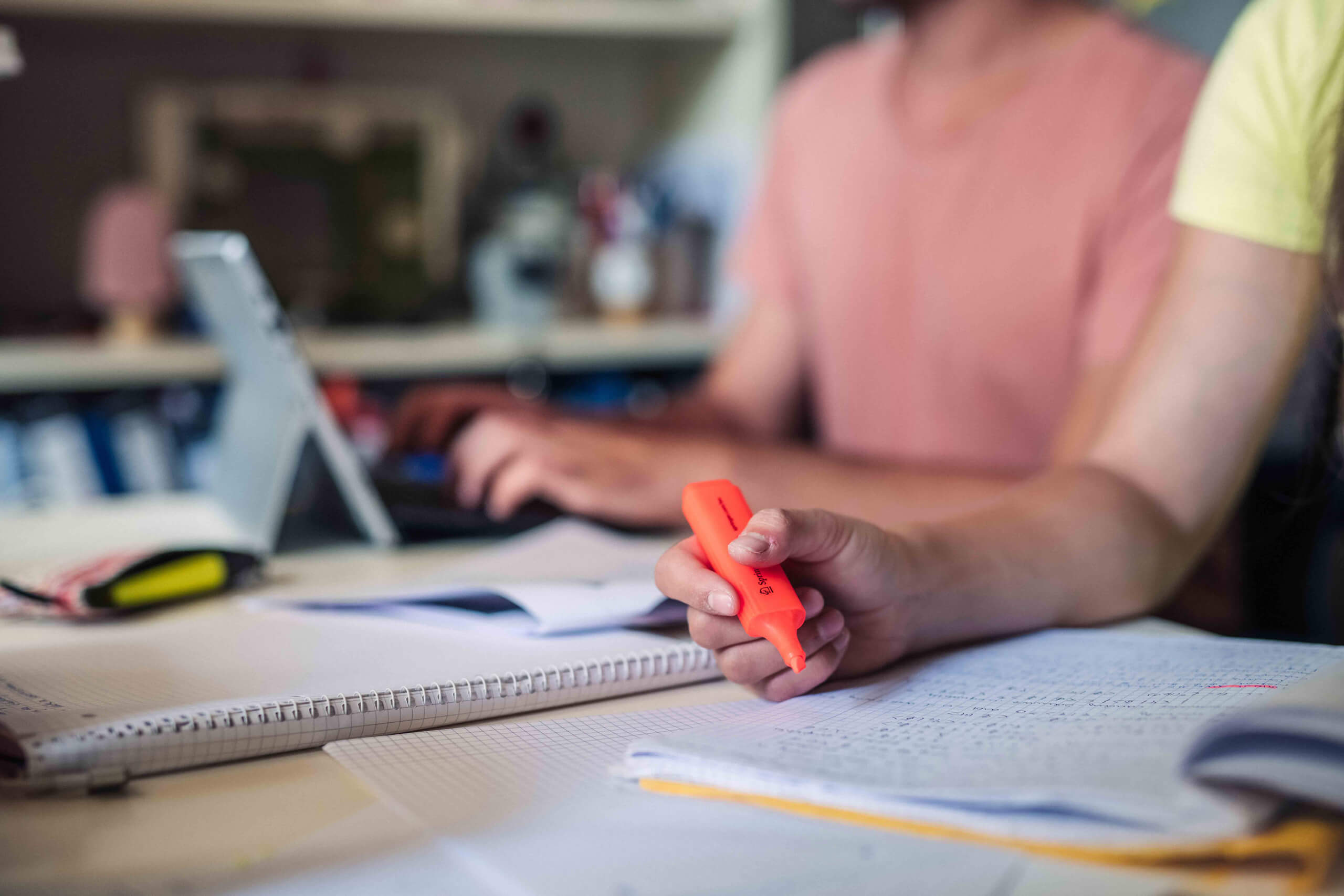 Eine Person hält einen orangefarbenen Textmarker über ein offenes Notizbuch voller Notizen, während eine andere Person im Hintergrund an einem Tablet arbeitet. Auf dem Arbeitsplatz liegen zahlreiche Papiere und Bücher verteilt.