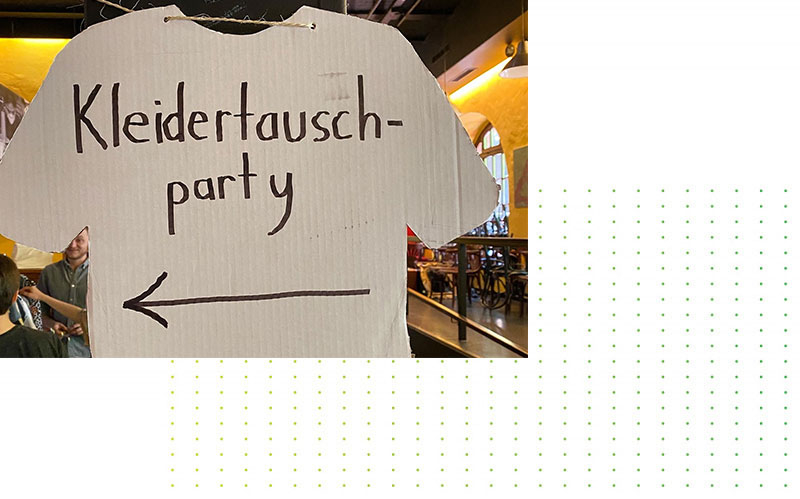 Von der Decke hängt ein Pappaufsteller in Form eines T-Shirts, auf dem handschriftlich „Kleidertausch-Party“ und ein nach links zeigender Pfeil stehen, was auf eine Kleidertausch-Veranstaltung in einem Café oder Restaurant hinweist.