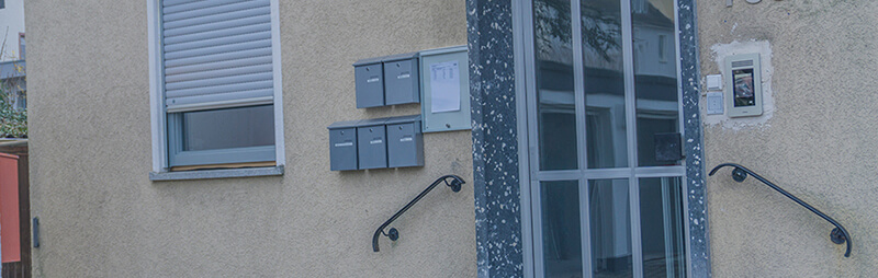 Eingang zu einem beigen Studentenwohnheim mit einer Glastür, fünf grauen Briefkästen, einem geschlossenen Fenster mit Rollladen und zwei schwarzen Handläufen neben Steinstufen.