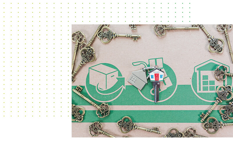 Eine dekorative Anordnung von alten Messing-Schlüsseln liegt auf einem Karton mit grünen Symbolen für Umzug, Transport und Lagerung. In der Mitte befindet sich ein Schlüsselbund mit einem Hausschlüssel und einem Anhänger in Form eines Hauses mit der Aufschrift „HOME“.