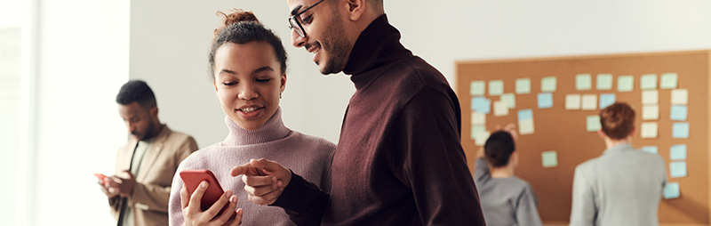 Zwei junge Erwachsene schauen gemeinsam auf ein Smartphone, im Hintergrund arbeiten Personen an einer Pinnwand.