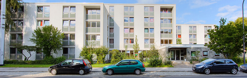 Modernes Wohngebäude mit heller Fassade, vielen Fenstern und bepflanztem Vorgartenbereich an der Straße