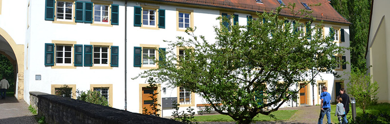 Ein historisches Gebäude mit weißer Fassade, grünen Fensterläden und rotem Dach; davor ein gepflasterter Weg und ein kleiner Baum im Innenhof.