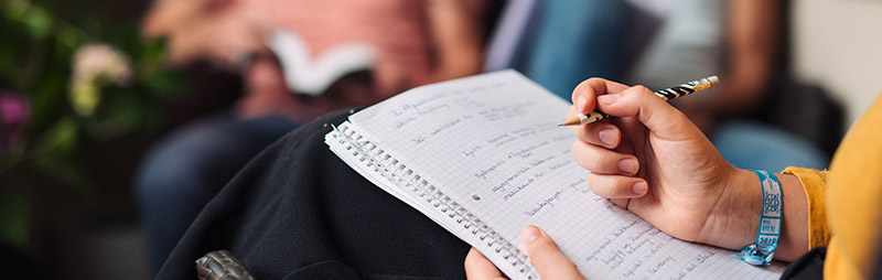 Eine Person schreibt mit einem Bleistift in ein kariertes Notizbuch, im Hintergrund unscharfe Menschen.