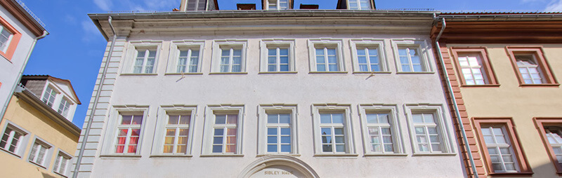 Historisches Wohnhaus mit symmetrischer Fensteranordnung, hellem Anstrich und geschwungenem Schriftzug über dem Eingang