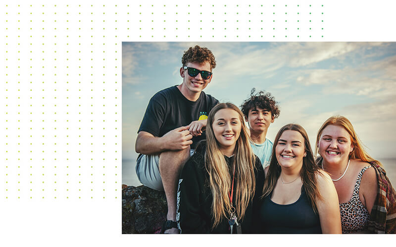 Fünf junge Menschen lächeln im Freien, im Hintergrund ein leicht bewölkter Himmel. Eine Person trägt eine Sonnenbrille und sitzt auf einem Felsen, die anderen stehen dicht beieinander und wirken alle glücklich und entspannt.