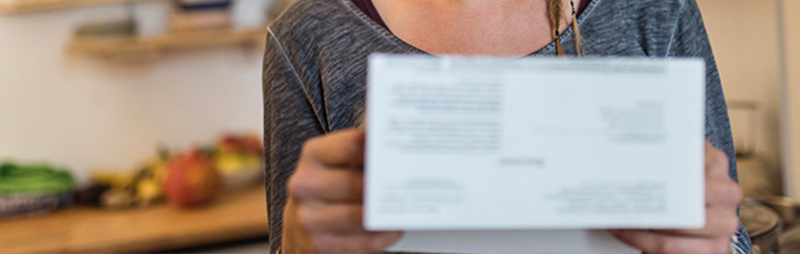 Eine Frau in einem grauen Hemd hält in einer Küche einen unscharfen Zettel in die Kamera. Im Hintergrund sind Obst und Regale zu sehen.