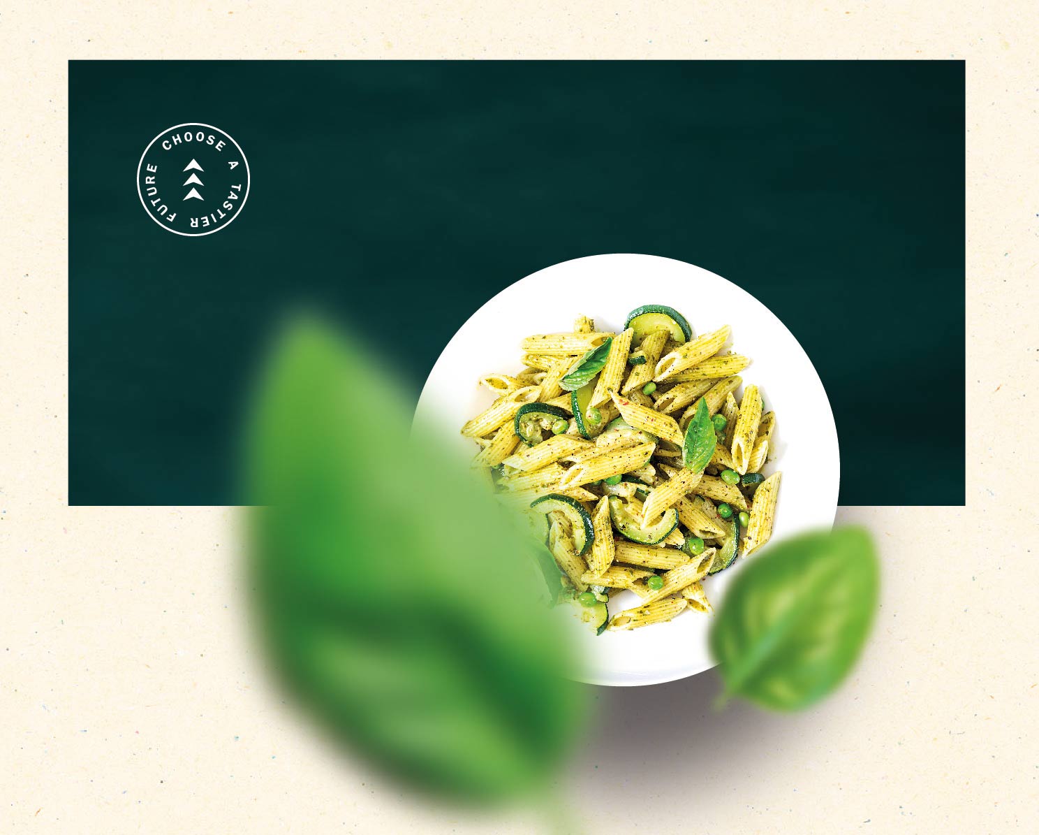Eine weiße Schüssel mit Penne-Nudeln, Zucchin und weiterem Gemüse in grüner Pesto-Sauce steht auf einem dunkelgrünen Hintergrund. Im Vordergrund sind zwei große, unscharfe Basilikumblätter zu sehen. In der Ecke befindet sich ein rundes Logo mit der Aufschrift „Choose a tastier future“.