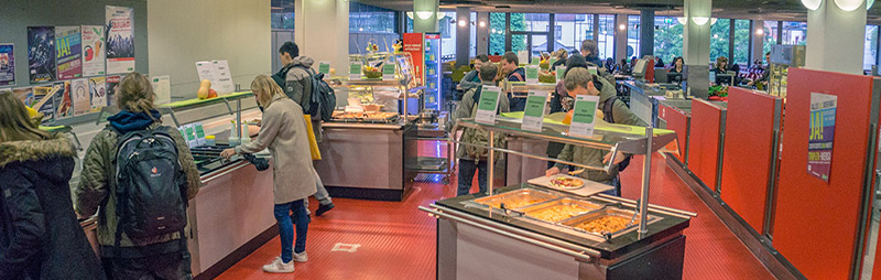 Studierende in einer modernen Hochschulmensa beim Mittagessen an Selbstbedienungstheken mit vielfältigem Speisenangebot.