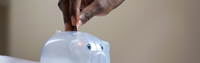 Eine Hand wirft eine Münze in ein durchsichtiges Sparschwein. Der Hintergrund ist weich verschwommen.