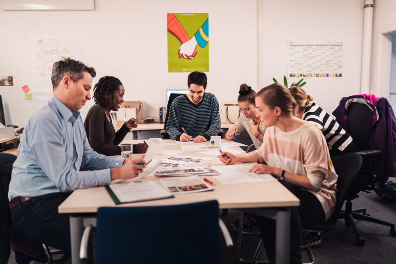 Gruppe von sechs Personen sitzt gemeinsam an einem Tisch in einem Büro und arbeitet konzentriert an Unterlagen