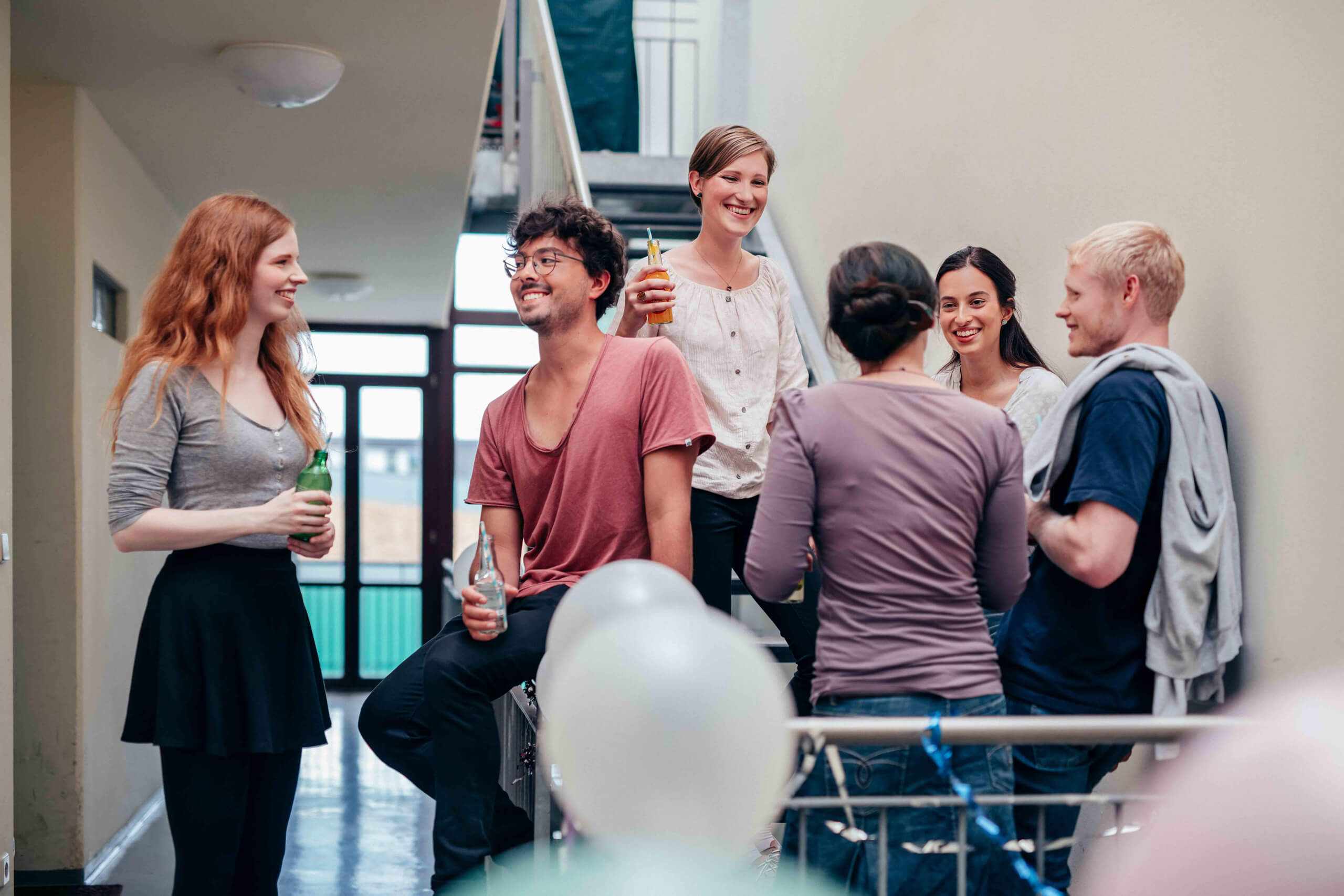 Mehrere Studierende feiern gemeinsam im Treppenhaus eines Wohnheims, sie unterhalten sich lachend mit Getränken in der Hand