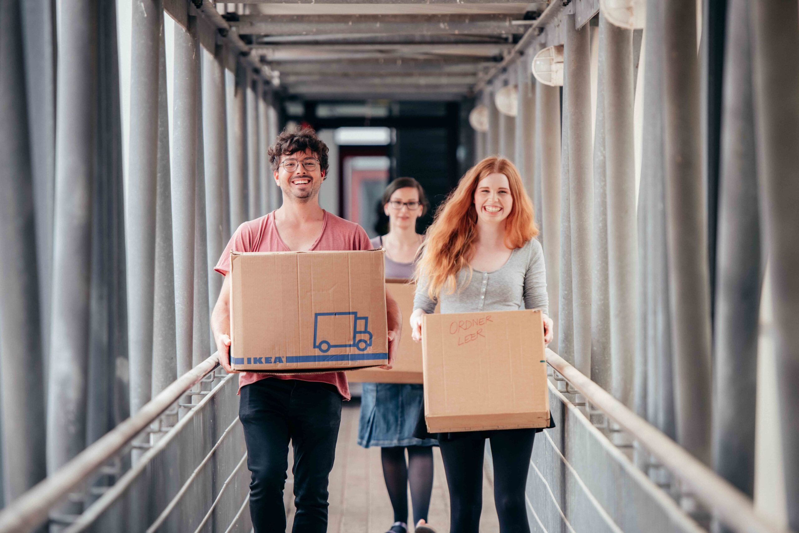 Drei Menschen gehen lächelnd und glücklich mit Pappkartons über eine Brücke. Entlang des Gehwegs sind Metallgeländer und -balken zu sehen.