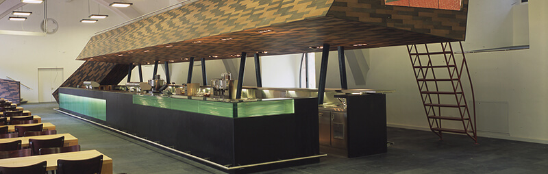 Innenaufnahme einer historischen Mensa mit moderner, schwarzer Theke, auf der Speisen angerichtet sind.