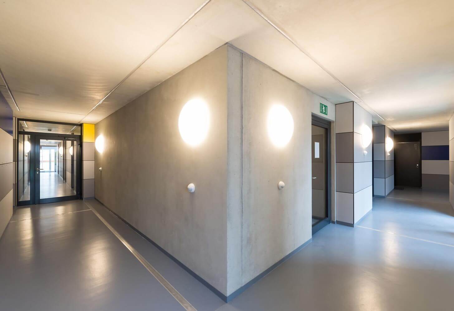 Moderner Flur mit klarer Architektur, hellen Wänden und markanten Wandlampen, die symmetrisch auf beiden Seiten eines Gebäudeecks angebracht sind.
