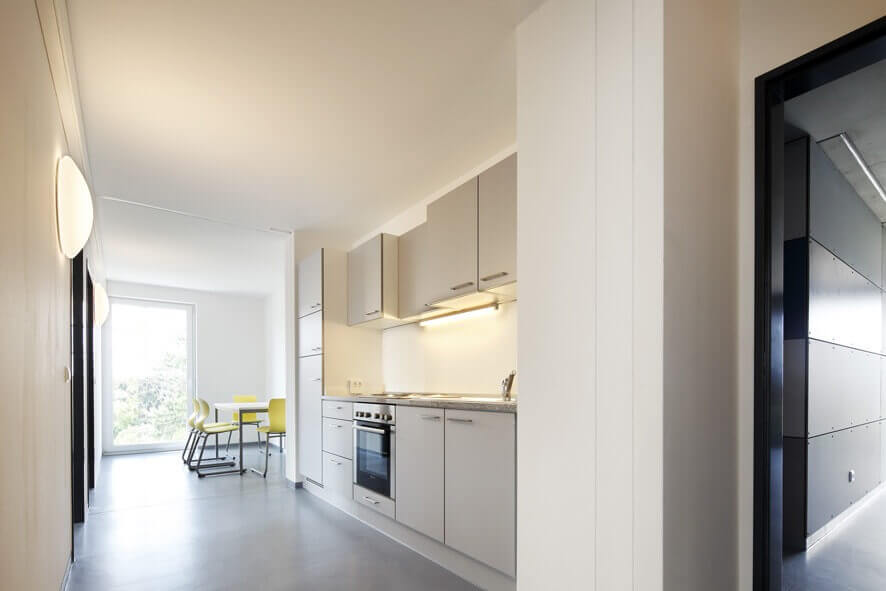 Gemeinschaftsküche mit integrierter Küchenzeile, gelben Stühlen und Esstisch, hellem Bodenbelag und freundlicher Beleuchtung, aufgenommen vom Eingangsbereich aus.