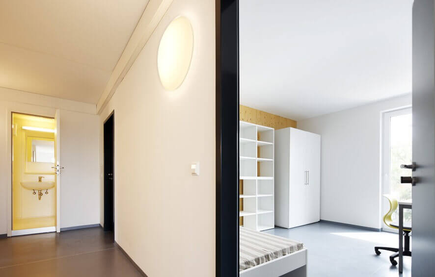 Offener Blick von einem Flur in ein modern eingerichtetes Wohnheimzimmer mit Bett, Kleiderschrank und Schreibtisch vor einem großen Fenster, gestaltet in Weiß und Gelb.