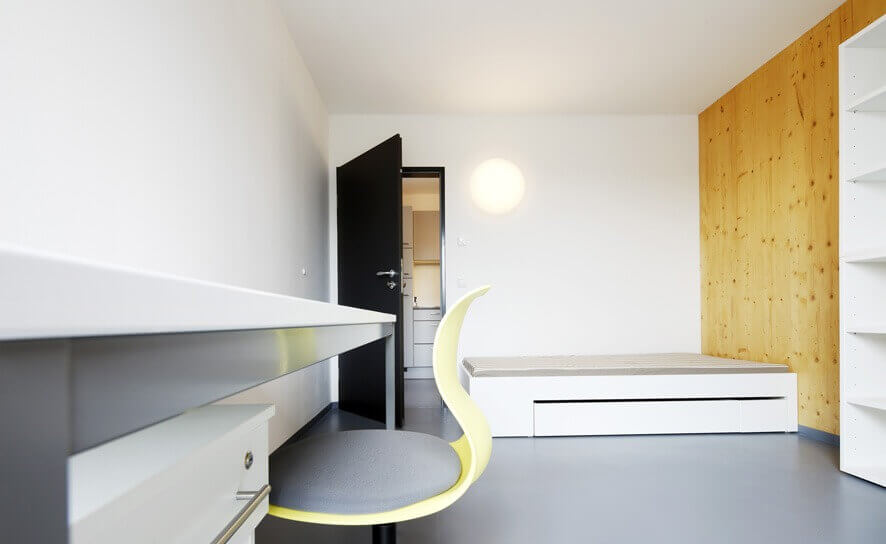 Blick aus einem Flur in ein Wohnheimzimmer mit Regal, Kleiderschrank, Bett und großer Fensterfront, modern möbliert mit hellen Farben und gelben Akzenten.