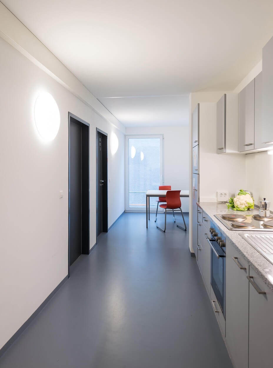 Gemeinschaftsküche mit Esstisch und zwei roten Stühlen, moderner Einbauküche und hellen Wänden, aufgenommen mit Blick auf die Küchenzeile und den Flur.