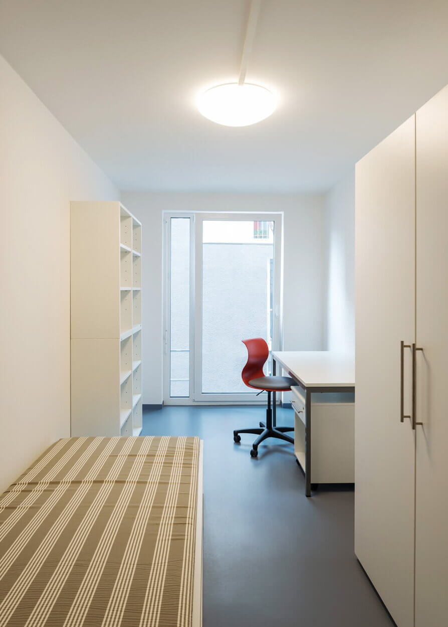 Modernes Einzelzimmer in einem Studentenwohnheim mit Bett, Schreibtisch, Regal und Kleiderschrank, klar strukturiert und hell beleuchtet, mit Blick auf die Fensterfront.